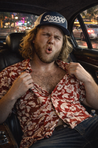 Man sitting in a car wearing a red floral shirt and a cap with 'SHODOO' branding.